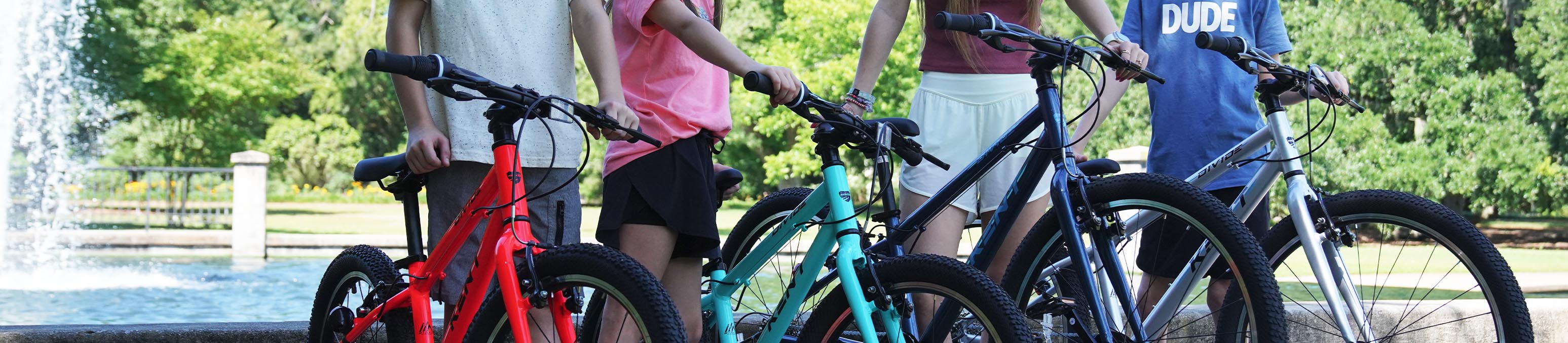 Kids with discount bikes standing by a fountain outdoors, holding colorful bicycles in a park.